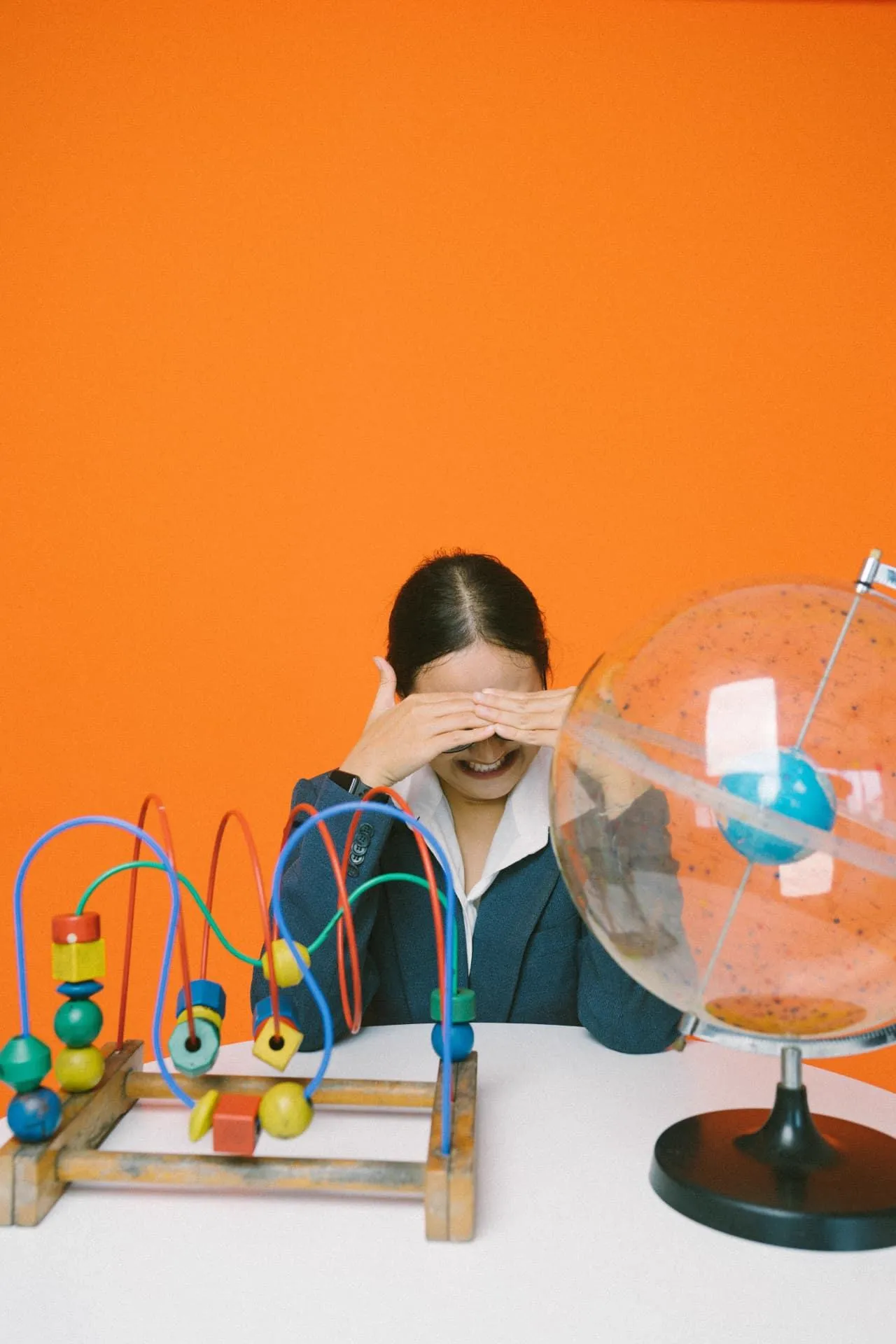 A woman playing peekaboo behind a table with a colourful bead toy and a globe, against a bright orange background
